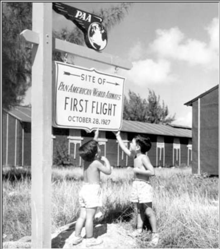 Children stand beneath a PAA sign marking the PAA's first international flight.